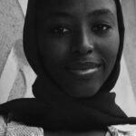 Documentaries - Black and white low angle of young smiling African American female in shawl on head and t shirt with text pattern standing leaned on concrete wall and looking at camera