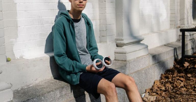 Virtual Fitness Coaches - Full body of young male in sportswear and VR goggles sitting on concrete foundation of white building and holding controllers
